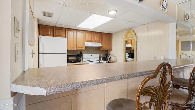 kitchen with a drop ceiling, a kitchen bar, white appliances, and kitchen peninsula