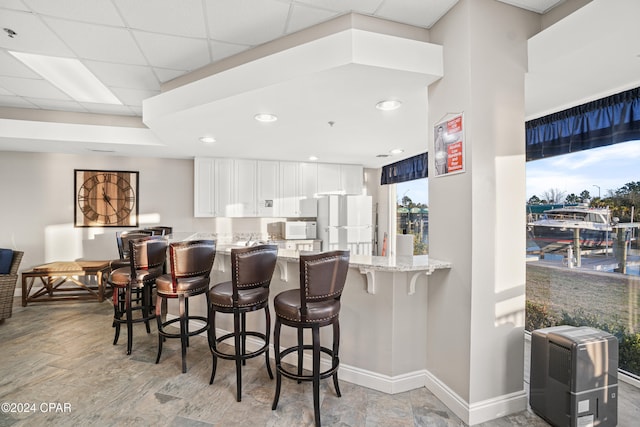 kitchen with a drop ceiling, white appliances, light stone counters, white cabinetry, and a breakfast bar area