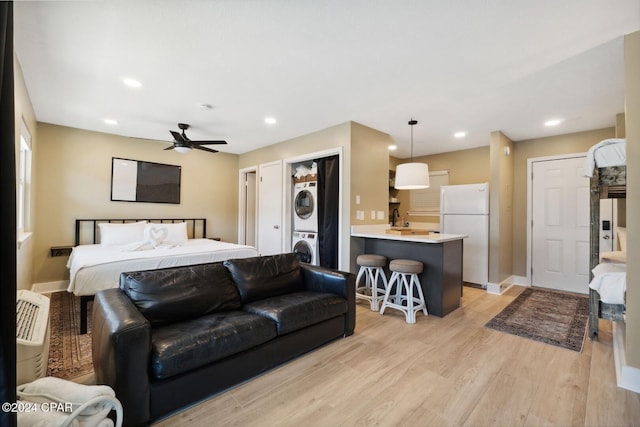 bedroom featuring light hardwood / wood-style flooring, ceiling fan, stacked washing maching and dryer, white fridge with ice dispenser, and white fridge