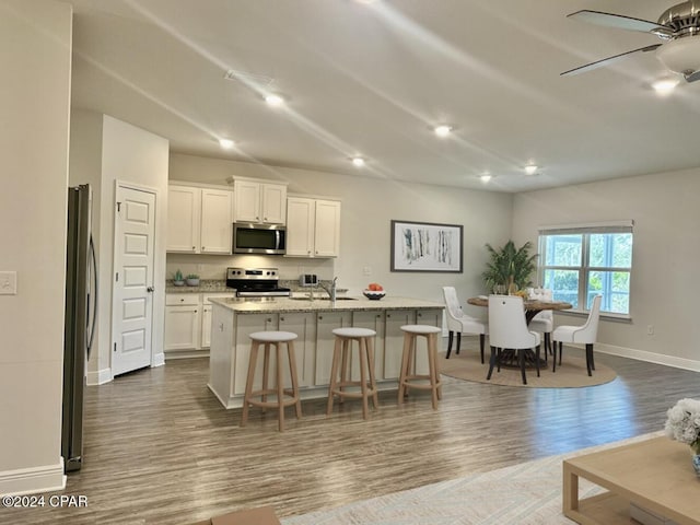 kitchen with light stone counters, wood-type flooring, a kitchen island with sink, white cabinets, and appliances with stainless steel finishes