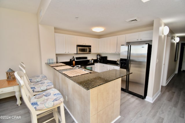 kitchen with white cabinetry, kitchen peninsula, dark stone countertops, light hardwood / wood-style floors, and appliances with stainless steel finishes