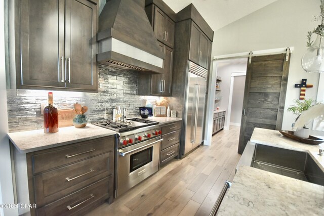 kitchen with high quality appliances, vaulted ceiling, a barn door, light stone counters, and custom range hood