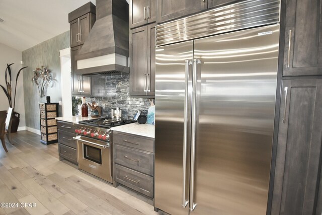 kitchen with high quality appliances, premium range hood, decorative backsplash, light wood-type flooring, and dark brown cabinetry