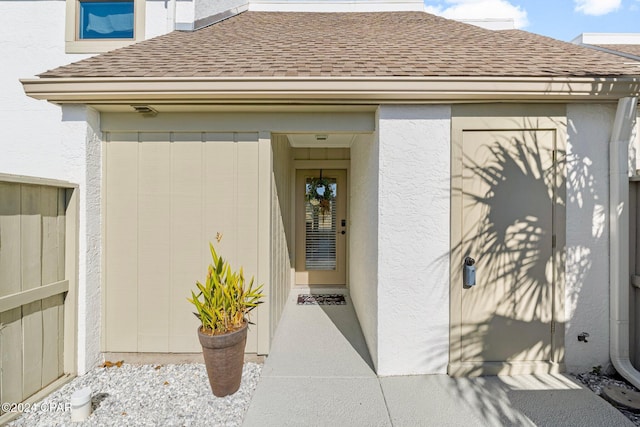 view of exterior entry featuring stucco siding, fence, and roof with shingles