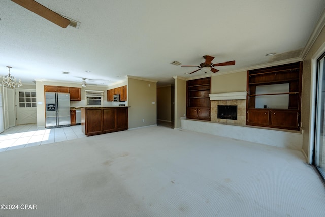 unfurnished living room featuring a tile fireplace, built in features, light colored carpet, ceiling fan with notable chandelier, and ornamental molding