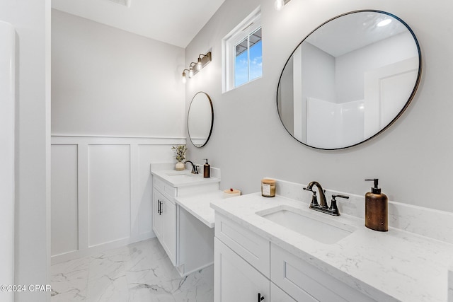 full bath with a sink, marble finish floor, two vanities, and a decorative wall