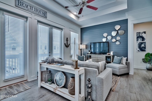 living room featuring ceiling fan, wood-type flooring, wooden walls, and a tray ceiling