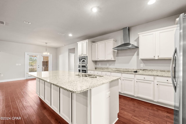 kitchen featuring appliances with stainless steel finishes, an island with sink, white cabinetry, wall chimney range hood, and decorative light fixtures