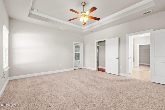 unfurnished bedroom with a spacious closet, a raised ceiling, light colored carpet, crown molding, and ceiling fan