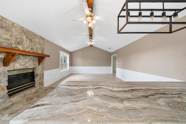 unfurnished living room with ceiling fan, a stone fireplace, lofted ceiling with beams, and light wood-type flooring