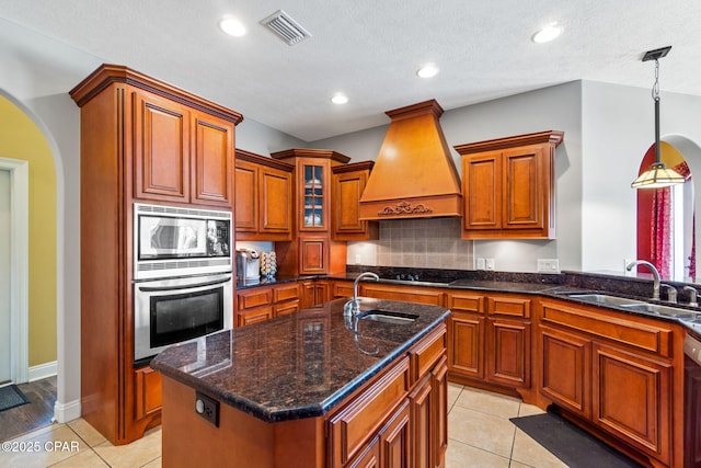 kitchen featuring premium range hood, oven, light tile patterned floors, an island with sink, and decorative light fixtures