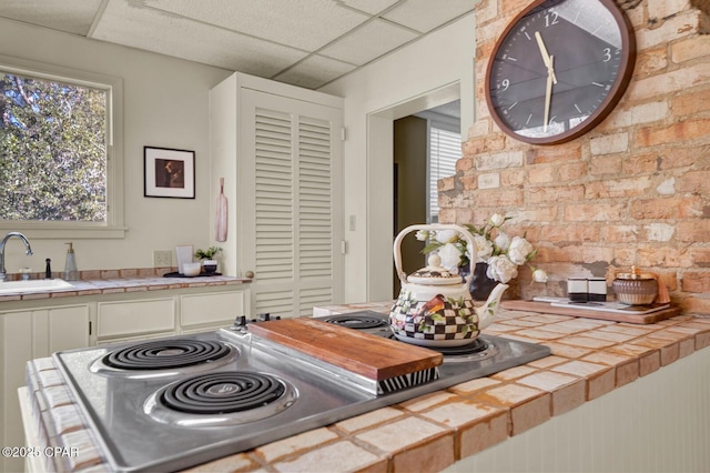 kitchen with tile counters, a paneled ceiling, sink, and brick wall