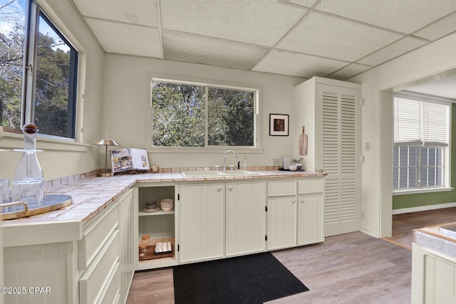 kitchen with a drop ceiling, sink, white cabinetry, light wood-type flooring, and tile counters