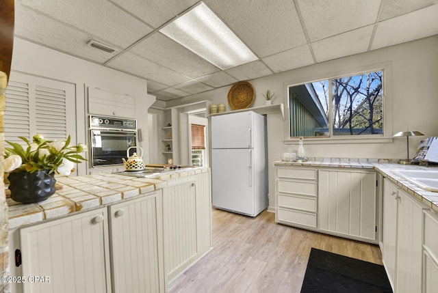 kitchen featuring tile countertops, oven, white fridge, white cabinetry, and a paneled ceiling