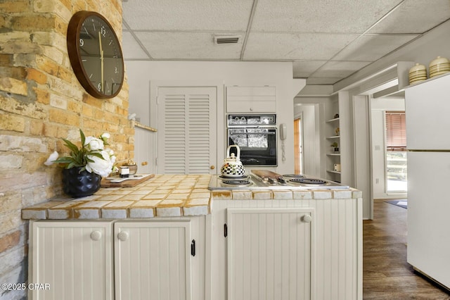 kitchen featuring tile counters, black oven, white cabinetry, white refrigerator, and a drop ceiling