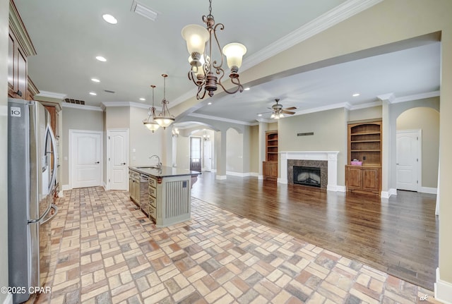 kitchen with sink, crown molding, hanging light fixtures, stainless steel refrigerator, and an island with sink