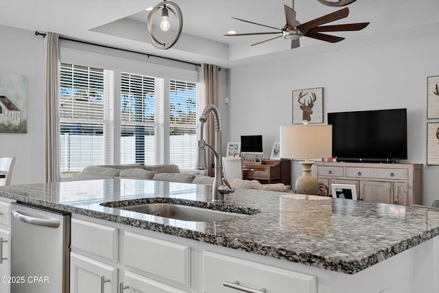 kitchen with a raised ceiling, dark stone countertops, stainless steel dishwasher, white cabinets, and sink