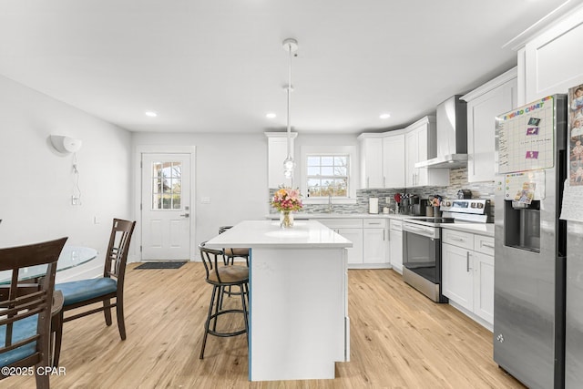 kitchen with wall chimney exhaust hood, hanging light fixtures, a kitchen island, stainless steel appliances, and white cabinets