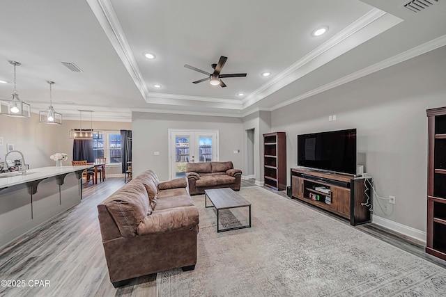 living room featuring sink, a tray ceiling, light wood-type flooring, ornamental molding, and ceiling fan with notable chandelier