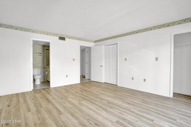 unfurnished bedroom with a textured ceiling, light hardwood / wood-style flooring, two closets, and ensuite bath