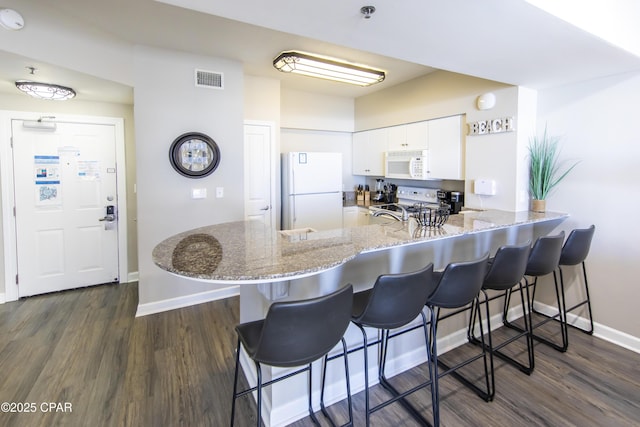 kitchen with kitchen peninsula, dark wood-type flooring, white cabinetry, white appliances, and a breakfast bar area