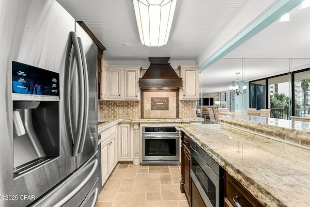 kitchen featuring premium range hood, cream cabinets, a notable chandelier, backsplash, and appliances with stainless steel finishes