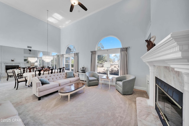 carpeted living room featuring a high ceiling, ornamental molding, ceiling fan with notable chandelier, and a high end fireplace