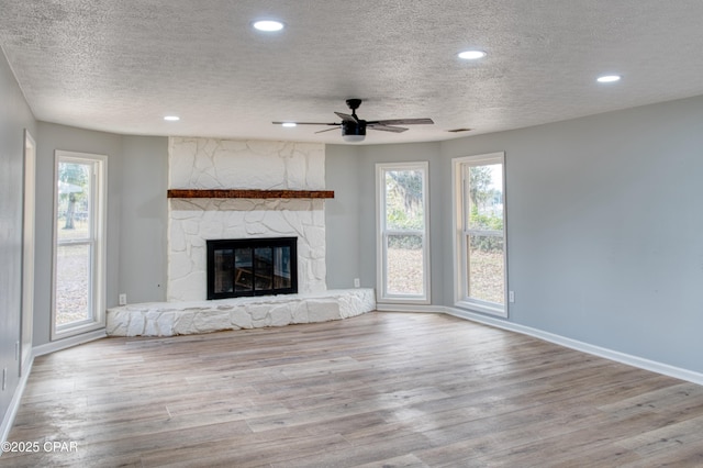 unfurnished living room with ceiling fan, a stone fireplace, a textured ceiling, and light hardwood / wood-style flooring