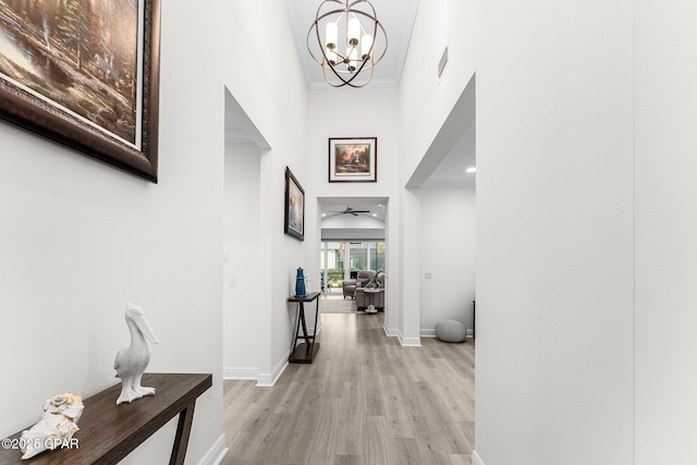 hall featuring light wood finished floors, baseboards, ornamental molding, a high ceiling, and a notable chandelier