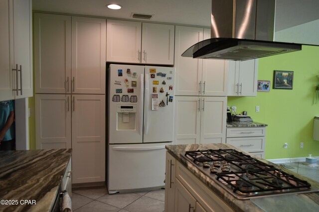 kitchen with light tile patterned flooring, island range hood, white cabinetry, white refrigerator with ice dispenser, and stainless steel gas cooktop