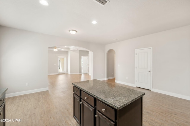 kitchen with dark brown cabinetry, light wood-type flooring, a kitchen island, ceiling fan, and dark stone counters