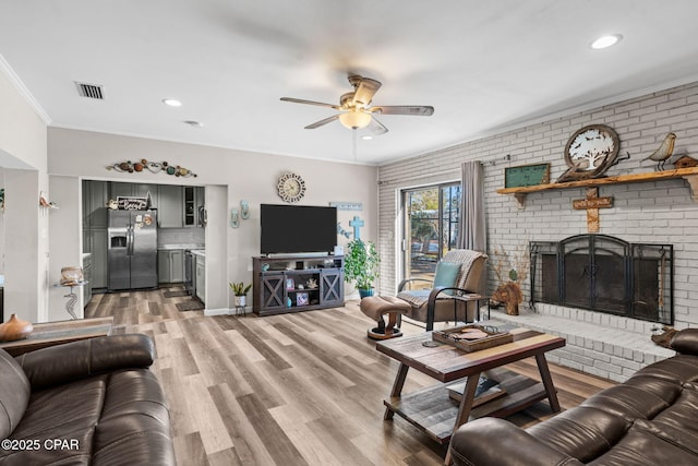 living room with ceiling fan, a brick fireplace, wood-type flooring, crown molding, and brick wall