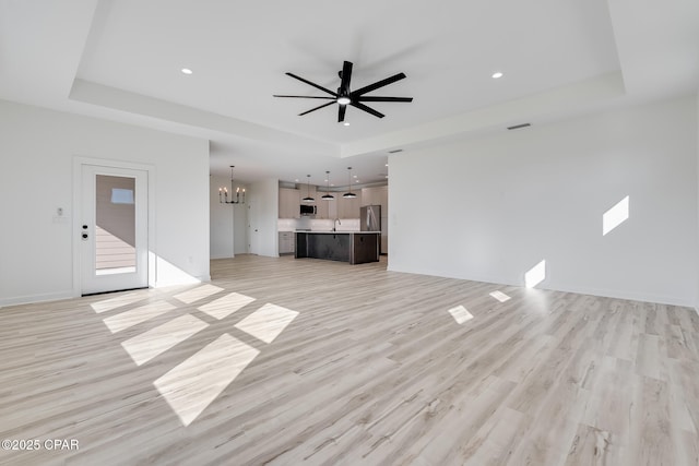 unfurnished living room with light wood-type flooring, ceiling fan with notable chandelier, and a raised ceiling