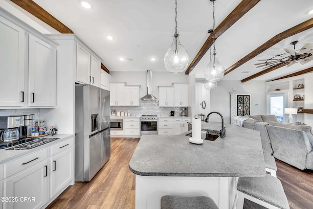 kitchen featuring sink, wall chimney range hood, stainless steel appliances, white cabinets, and beamed ceiling