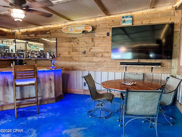 dining room with a bar, concrete flooring, ceiling fan, and wood walls