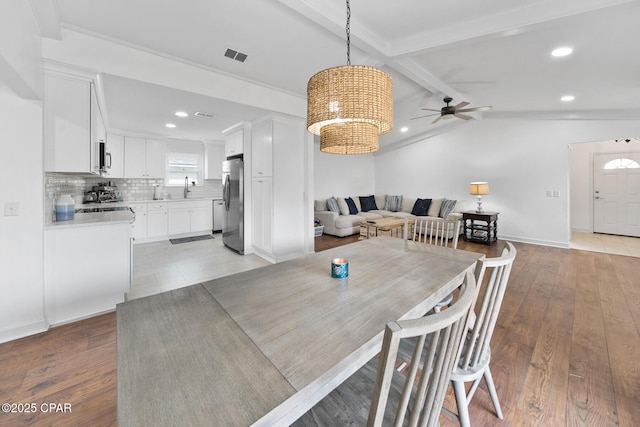 dining room with ceiling fan with notable chandelier, sink, lofted ceiling with beams, and light wood-type flooring