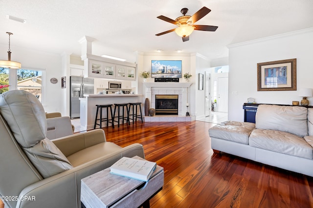 living room featuring a tiled fireplace, crown molding, dark wood-type flooring, and ceiling fan