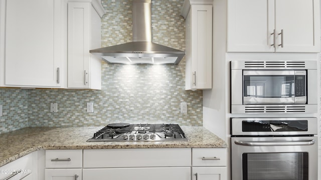 kitchen featuring white cabinetry, light stone counters, stainless steel appliances, decorative backsplash, and wall chimney range hood