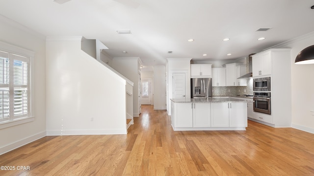 kitchen featuring light stone counters, appliances with stainless steel finishes, an island with sink, wall chimney range hood, and white cabinets