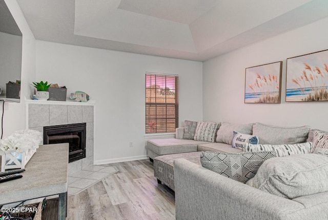 living room featuring a tiled fireplace, a raised ceiling, baseboards, and light wood finished floors