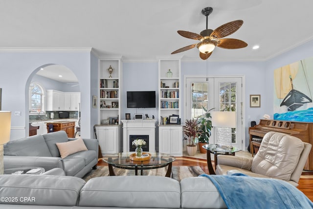 living room featuring arched walkways, a fireplace, a ceiling fan, light wood-style floors, and crown molding