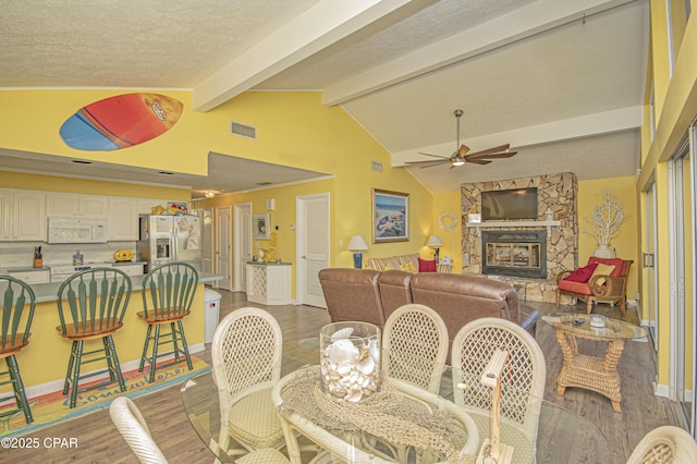 dining space featuring a fireplace, vaulted ceiling with beams, ceiling fan, light hardwood / wood-style floors, and a textured ceiling