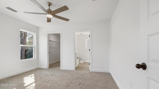 unfurnished bedroom featuring connected bathroom, a walk in closet, light colored carpet, a closet, and ceiling fan