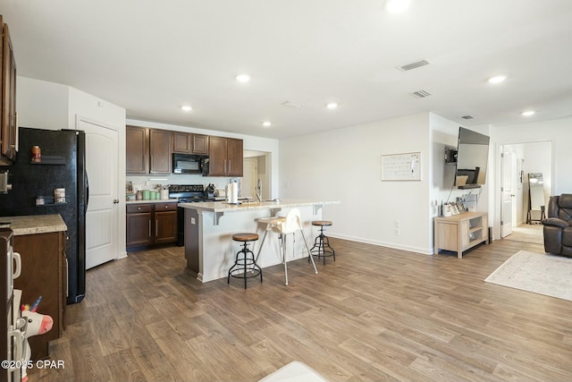 kitchen with wood-type flooring, a breakfast bar area, dark brown cabinetry, black appliances, and a center island with sink