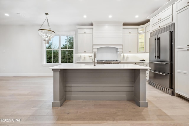 kitchen featuring high quality fridge, light countertops, wall chimney exhaust hood, an island with sink, and glass insert cabinets