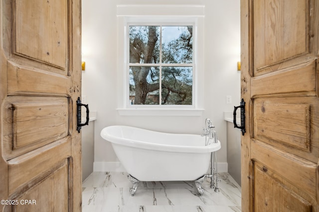 bathroom featuring marble finish floor, a healthy amount of sunlight, a freestanding bath, and baseboards