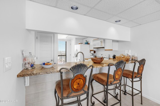 kitchen featuring a breakfast bar, white cabinetry, a paneled ceiling, kitchen peninsula, and white appliances