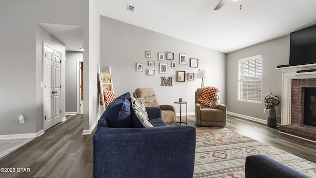 living room with a ceiling fan, visible vents, baseboards, a brick fireplace, and dark wood finished floors