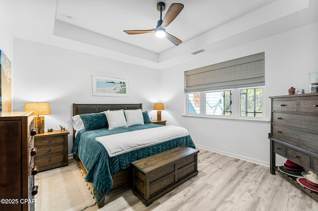 bedroom featuring visible vents, baseboards, light wood-type flooring, a tray ceiling, and a ceiling fan