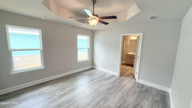 unfurnished bedroom featuring ceiling fan, ensuite bathroom, a raised ceiling, and light hardwood / wood-style flooring